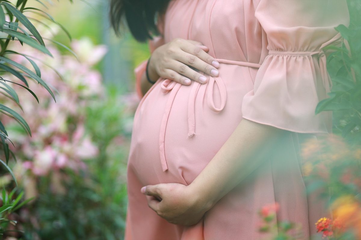 Mulher grávida em uniforme de trabalho doméstico na cozinha de uma casa, com a mão na barriga
