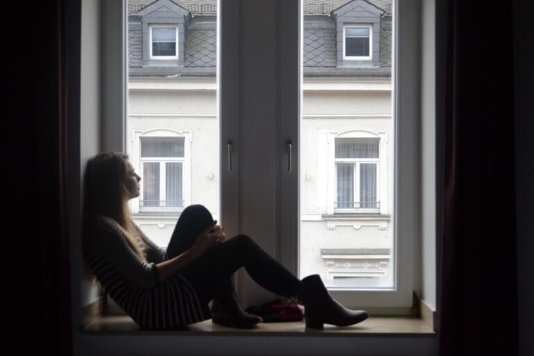 Mulher sentada perto da janela, com expressão pensativa, em casa
