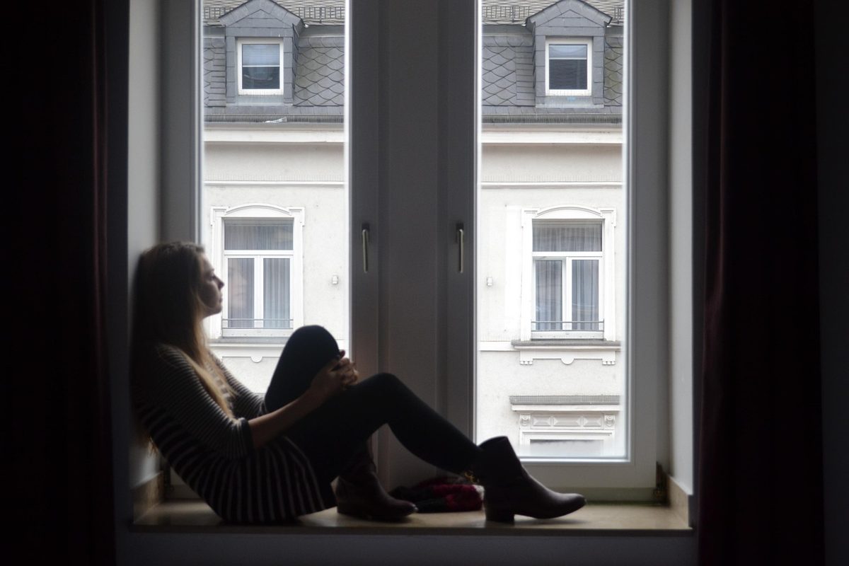 Mulher sentada perto da janela, com expressão pensativa, em casa